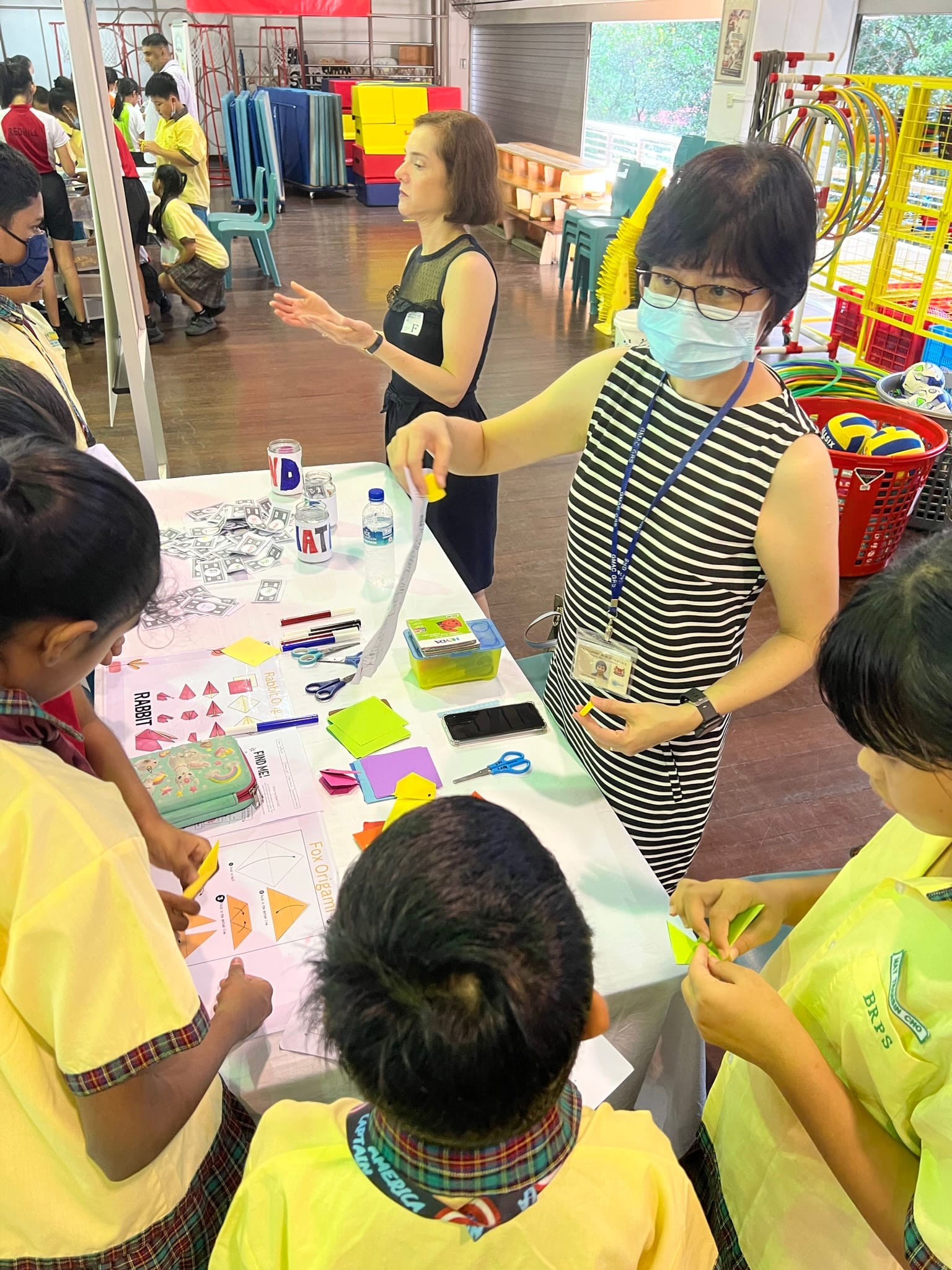Two teachers guide young students in an origami activity at a table covered with paper, scissors, and instruction sheets.